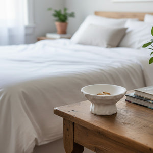 A small Luna Pedestal Dish Alba Lunarie with a textured, wavy rim sits on a rustic wooden bedside table in a softly lit bedroom. The background shows a neatly made bed with white linens and pillows, with a blurred plant on a nightstand.