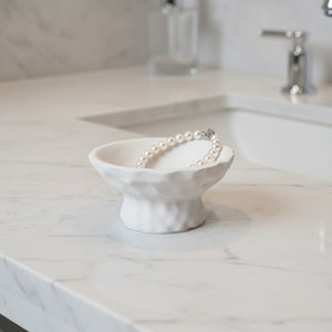 A white textured Luna Pedestal Dish Alba Lunarie jewelry stand holding a pearl bracelet, set on a white marble bathroom countertop near a sink.