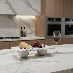 A modern kitchen scene with a white marble serving board on a marble countertop, styled with a wedge of cheese, red grapes, and a wine glass filled with red wine. The background shows light wood cabinets, stainless steel ovens, and a matching marble backsplash.