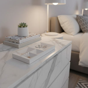 A white marble nightstand in a modern bedroom setting, styled with a small potted succulent, books, and a white compartment jewelry tray holding rings and a necklace.