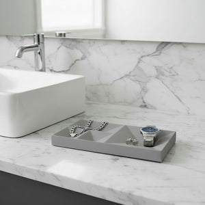 A rectangular gray jewelry organizer tray with geometric triangular compartments sits on a white marble bathroom counter next to a white vessel sink. The tray holds silver chain earrings and a silver watch.