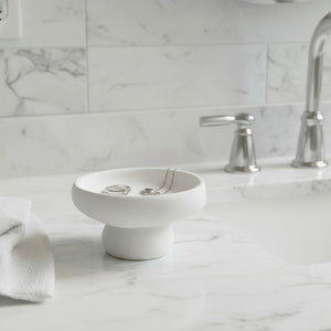 A Solene Pedestal Bowl  white stand with a shallow bowl on a pedestal base, placed on a white marble bathroom counter near a sink and faucet.