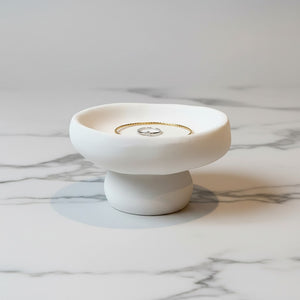 A Solene Pedestal Bowl  lunarie white jewelry display stand with a tiered base, holding a gold ring and a silver ring on a white marble surface.