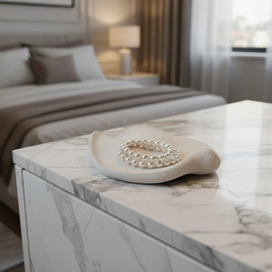 A cream colored textured jewelry organizer tray holding a pearl bracelet, set on a white marble-topped dresser in a softly blurred bedroom setting.