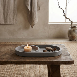 Eclipse Orbit Tray by Lunarie featuring a minimalist dual-circle design, ideal as a decorative catchall tray for jewelry and modern home décor. Gray tray with a lit candle and stones on a wooden table in a room with a window and vase.